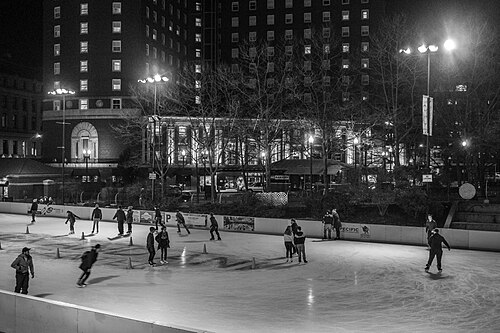 Skaters on the Alex and Ani City Center rink at Kennedy Plaza at night, a colonnaded stone building lit up behind bare trees.