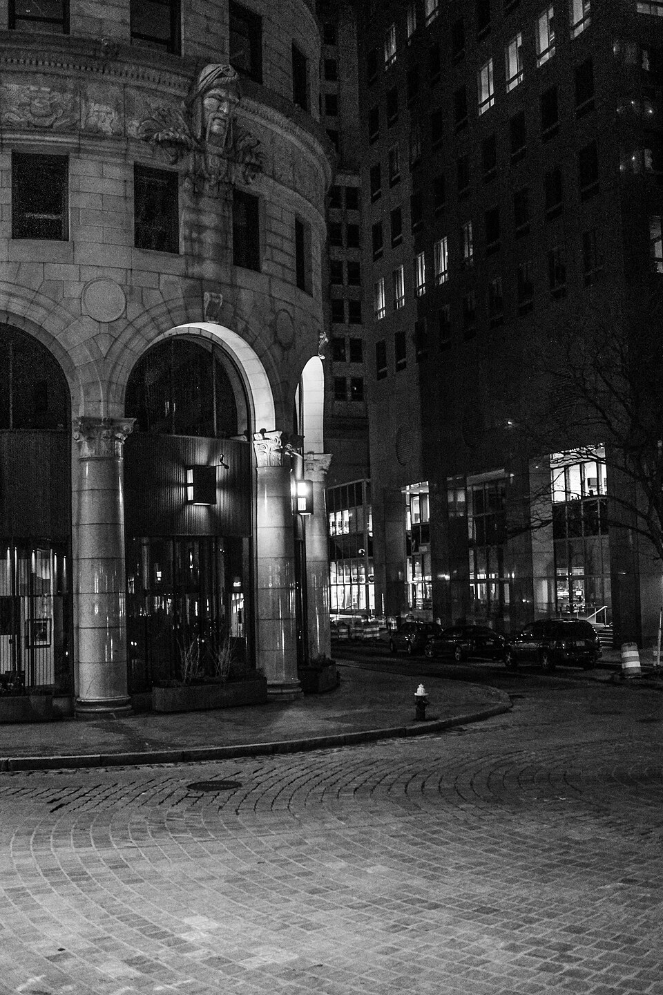 The rounded stone corner of the Turk's Head Building at night, a carved face under the cornice, paired columns over the arched entrances, and a brick and cobblestone plaza in front with a fire hydrant and street lamps.