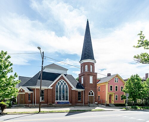 A red brick church with a tall dark spire beside a two storey pink clapboard house on Dexter Street.