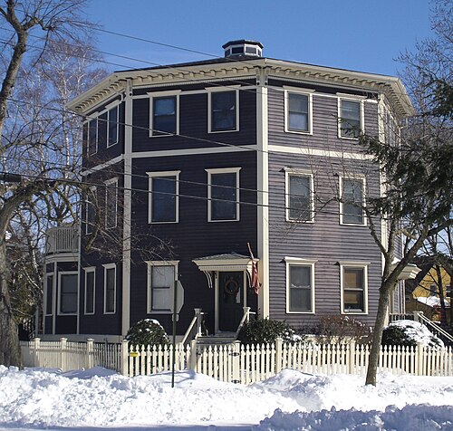 An octagonal dark clapboard house on Harrison Street behind a white picket fence.