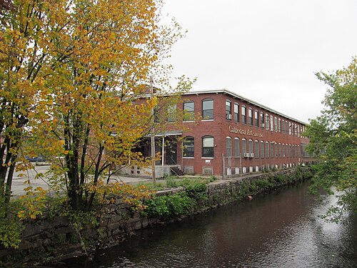 A long two storey red brick mill building alongside a narrow river channel bordered by stone walls.