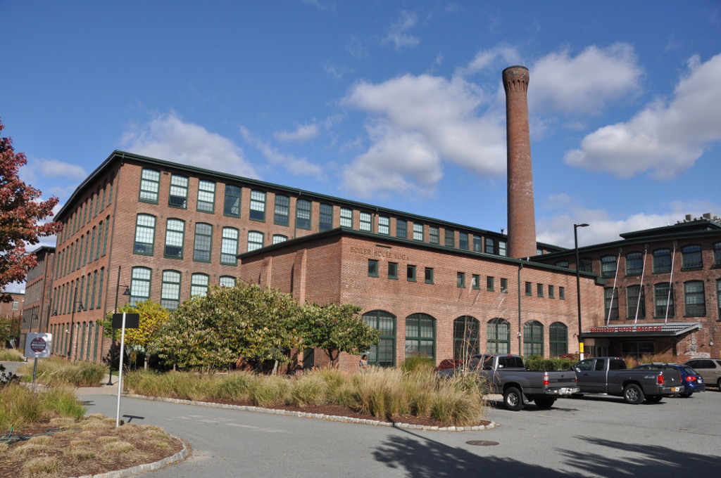 A large red brick mill complex with a tall brick smokestack beside an asphalt parking area.