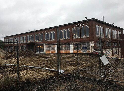 A two storey brick industrial mill building behind a chain link fence, disturbed dirt in the foreground.