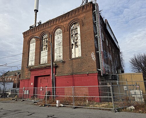 A tall brick utility building with arched window openings behind a chain link fence.