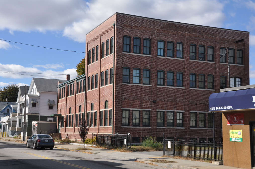 A three storey red brick commercial building on a corner next to residential houses along an asphalt street.