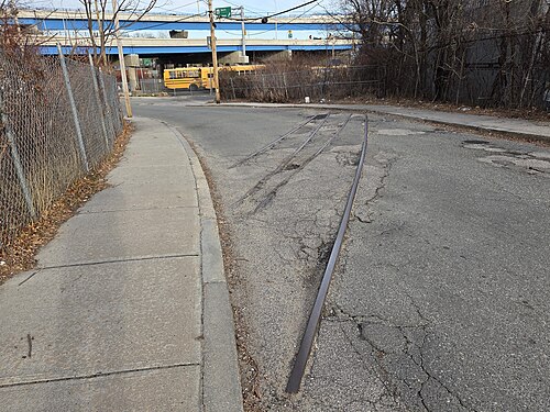An asphalt road with disused railroad tracks embedded in it, curving alongside a concrete sidewalk under an overpass.