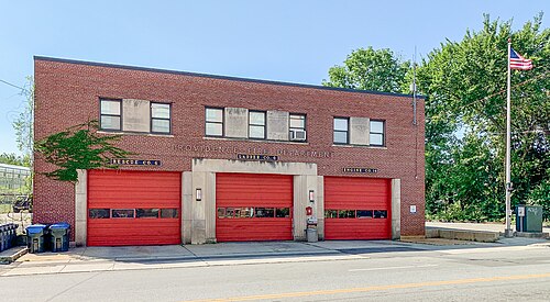 A two storey red brick fire station with three red garage doors and lettering above the entrances.