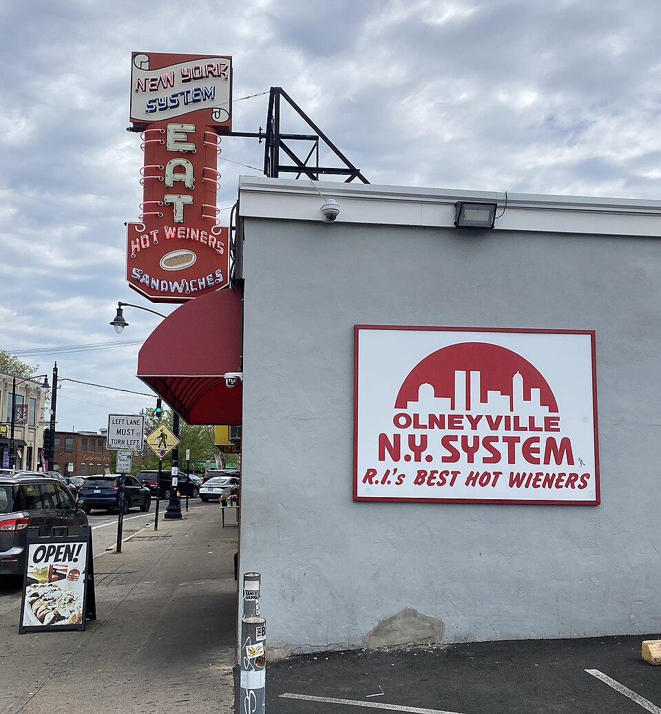 A grey painted commercial building exterior with a large white and red wall sign and a vertical neon roof sign.