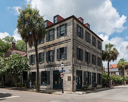 A three storey weathered grey stucco corner building with red mansard dormers, dark shutters, a ground floor shop and a gas lamp post, palmettos either side.