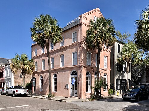 A three storey salmon pink stucco corner building with arched ground floor windows and a shallow parapet roof, palmettos and a pickup truck at the kerb.