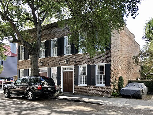 A two storey red brick house with black shutters and a white painted door surround, a single tree and a parked car in front.