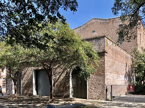 A brick warehouse corner with two arched doorways and a roll up garage door, a for sale sign on the fence and trees shading the sidewalk.