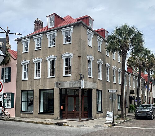 A three storey tan stucco corner building with white window trim, a standing seam red roof with two dormers, ground floor storefront glass and a one way sign at the corner.