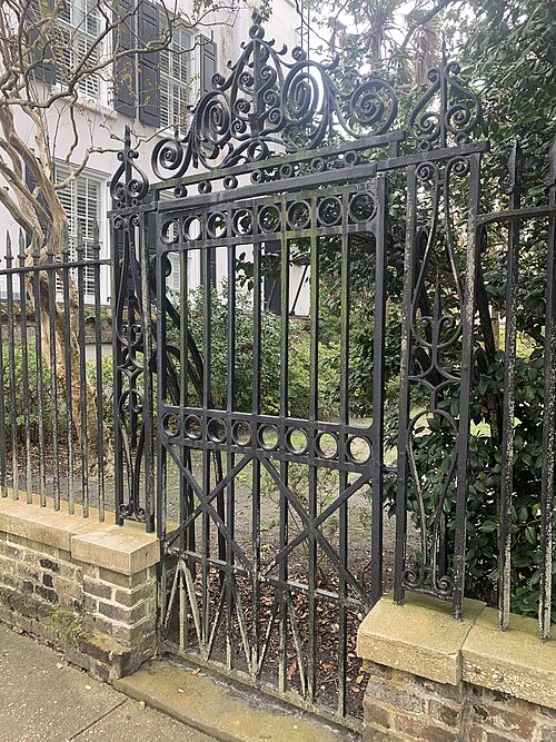 An ornate black wrought iron gate set into a low brick wall on Chalmers Street, opening onto Washington Square, with a white clapboard house visible through the greenery behind it.