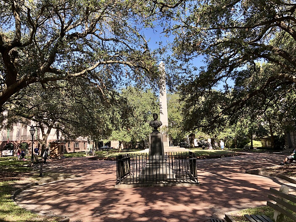 A brick paved park square under a canopy of mature live oaks, the granite Washington Light Infantry obelisk and bronze bust inside a low iron fence, a gas lamp post and pastel townhouses visible through the trees.