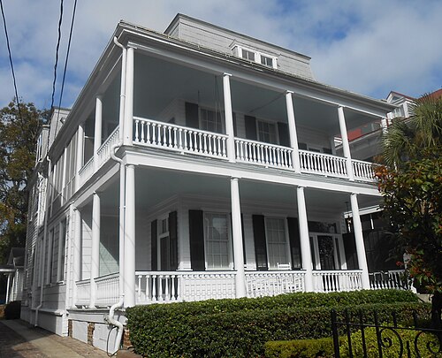A white wooden single house with two stacked tiered piazzas and black shutters, a garden path along its side.