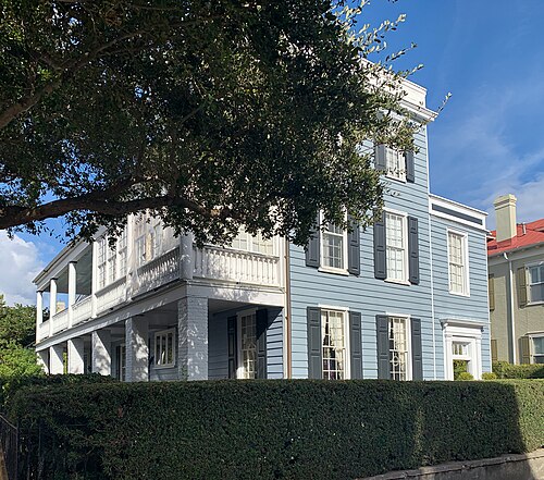 A pale blue single house turned side on to the street, a white tiered piazza under a live oak, behind a tall hedge.