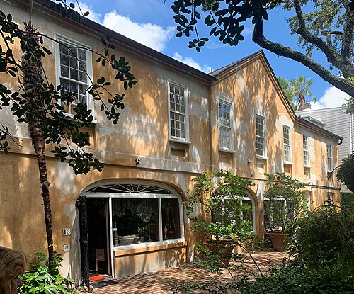 A yellow stucco carriage house with arched ground floor openings, opening onto a brick paved courtyard under live oaks.