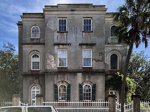 A weathered grey stone mansion with arched windows and green shutters, stucco peeling to bare brick, a palm tree at the fence.