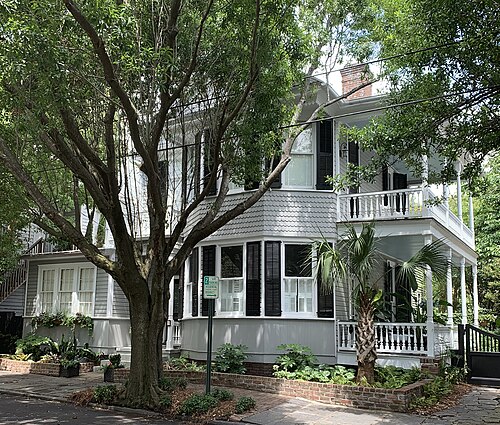 A grey shingled single house with black shutters and a bay window, its tiered porch behind a large street tree.
