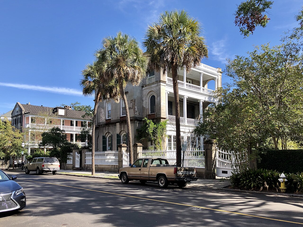 A sunny stretch of Meeting Street, single houses with tiered piazzas behind palm trees and pickup trucks parked at the kerb.