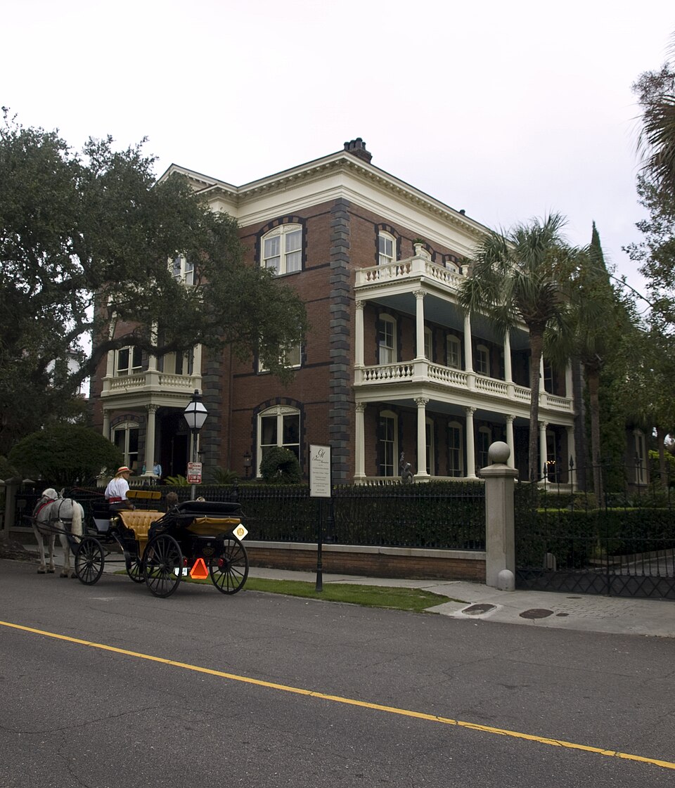 The Calhoun Mansion's brick and stone Italianate front, two stacked columned piazzas, palmettos at the fence line and a horse drawn carriage waiting at the kerb.