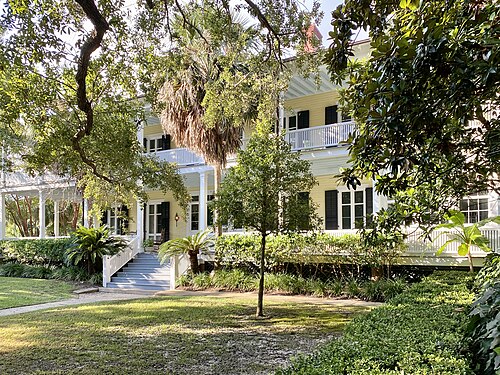A pale yellow two storey officer's house with double porches and a rooftop deck, oak branches overhanging the roofline.