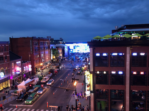 Lower Broadway at dusk from a rooftop bar on the 300 block, neon down both sides of the street and a temporary stage being built at the far end.