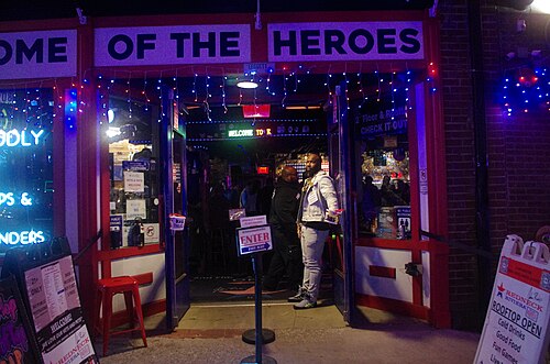The door of the Redneck Riviera at 208 Broadway at night, a doorman inside the open doors and the words HOME OF THE HEROES across the front above him.
