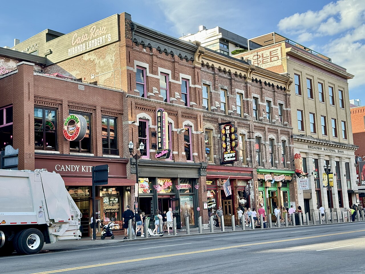 The north side of the 300 block of Broadway on a sunny afternoon, brick and stone fronts of three and four storeys with a sign on every one, a candy shop and a boot store at street level.