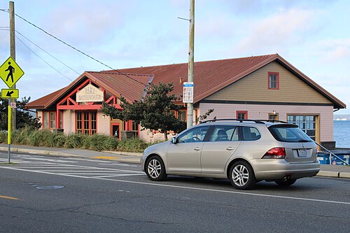 The one storey Alki Bathhouse on Alki Avenue SW, pink wood siding with red trim and a red tile roof, a car parked at the curb.