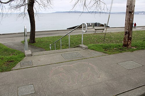 Concrete steps and a metal handrail lead down from the promenade to the beach, a bench and a painted animal outline on the paving.