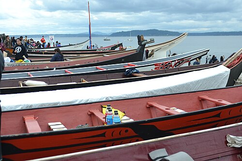 Carved wood canoes pulled up on the open beach at Alki, painted hulls and bow figures, people gathered along the waterline.