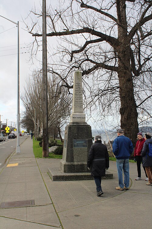 The sidewalk along Alki Avenue SW, overhead wires and street lamps, parked cars and low buildings across the road, a stone marker on the promenade.