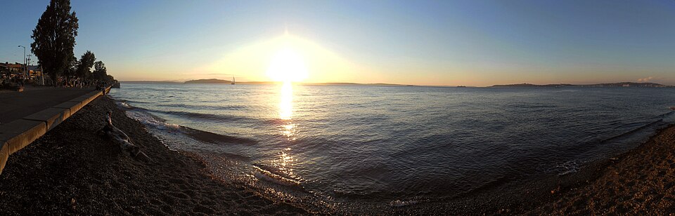 A stretch of open gravel beach and the concrete seawall promenade at Alki, waves breaking at the shoreline with the far side of the bay in the distance.