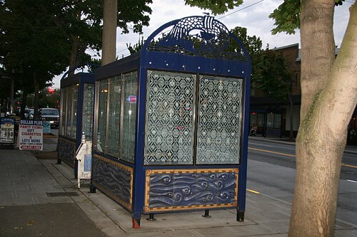 A blue tiled and glass transit shelter on the sidewalk at NW Market Street, a Dollar Plus storefront and brick building behind it.