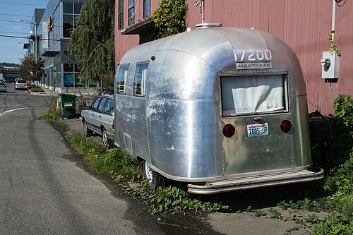 A vintage Airstream trailer parked beside a red corrugated metal industrial building on a back street in Ballard.