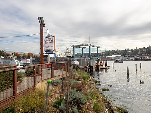 The Canal Boatyard sign and a travel lift over the water, work boats moored at the dock and the far shore of Salmon Bay behind.