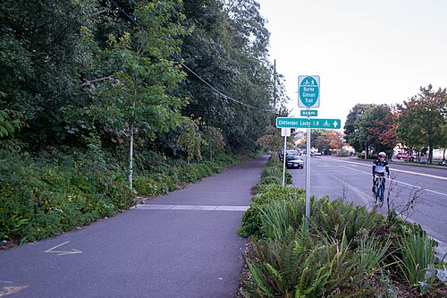A paved trail beside the road with a sign pointing toward the Chittenden Locks, a cyclist passing on the roadway alongside.