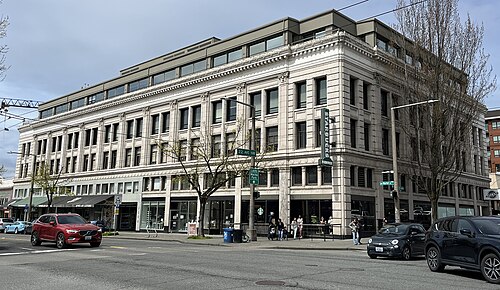 A stone faced corner building on Northwest Market Street, storefronts along the ground floor and cars parked at the curb.