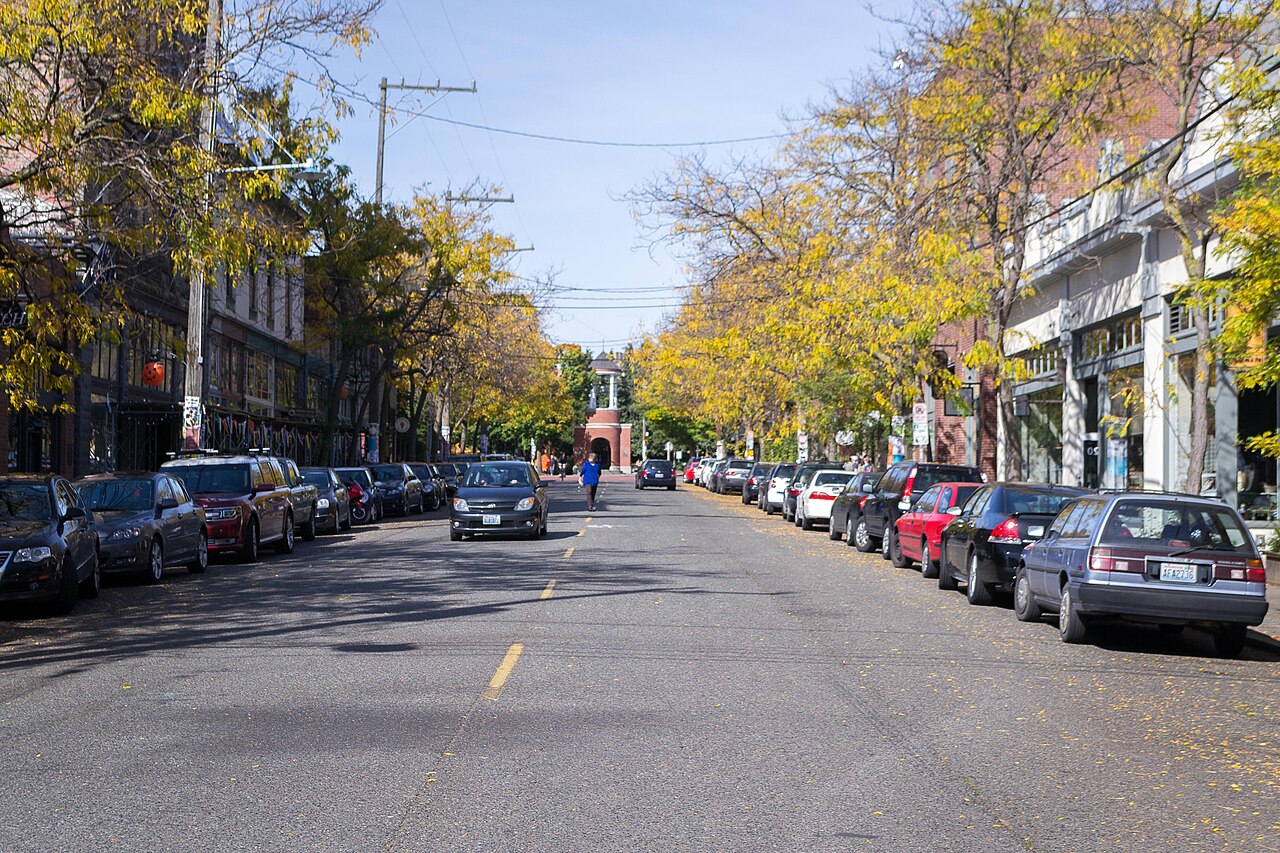 Ballard Avenue looking down the block, two and three storey brick storefronts on both sides, street trees overhead and cars parked along the curb.