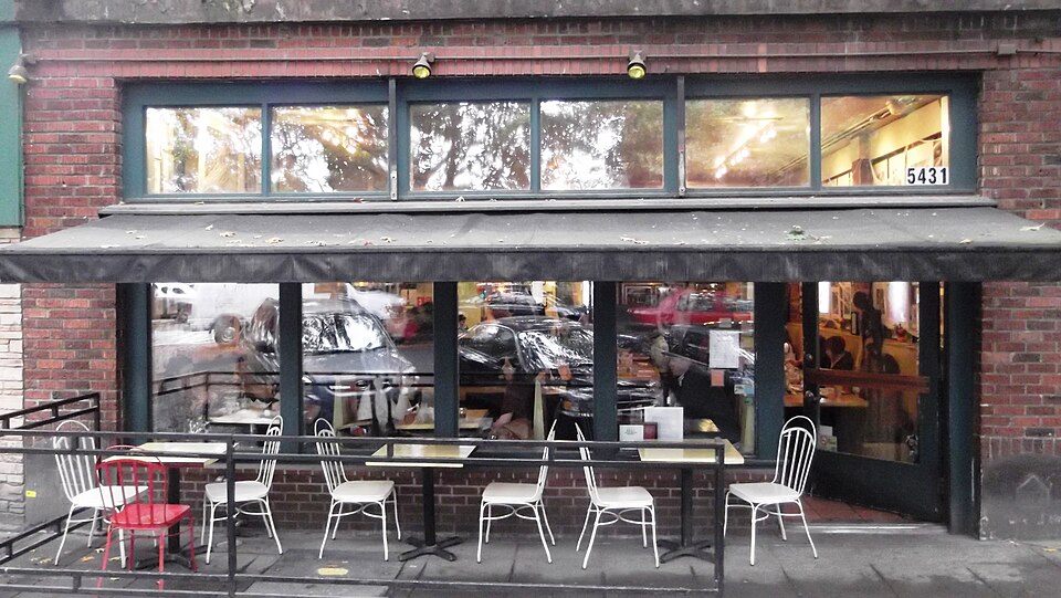 A brick storefront under a dark canvas awning at 5431 Ballard Avenue Northwest, white cafe chairs out front. The kind of Ballard Avenue storefront the florist shop occupied, not the building itself, which stands about 165 meters north.