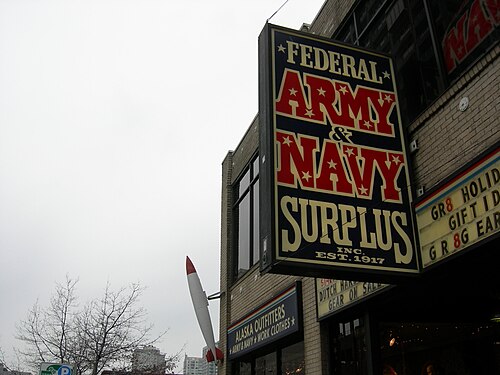 The Federal Army and Navy Surplus sign projects over 1st Avenue in red white and blue lettering, a brick storefront and an arched window below, a red and white finned sign shaped like a rocket mounted beside it as part of the store's own signage.