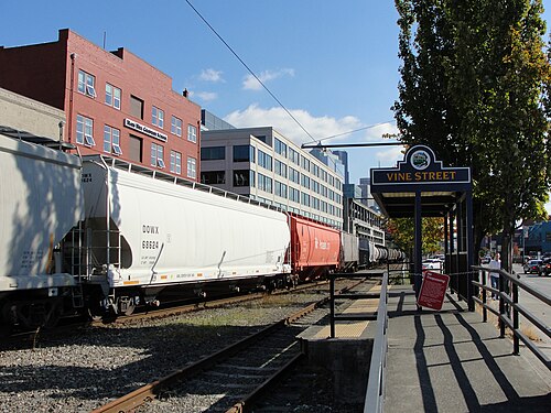 A red brick building with arched window heads stands beside the BNSF rail line at Vine Street, freight cars on the tracks and the old Vine Street streetcar shelter along the platform.