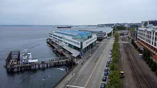 An elevated view down Alaskan Way, a pier and passenger terminal on the left, a double track rail line and parked cars along the roadway, downtown towers in the distance.