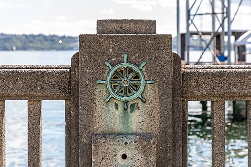 A ship's wheel medallion set into a concrete post along an Alaskan Way pier railing, the water and a dock behind it.