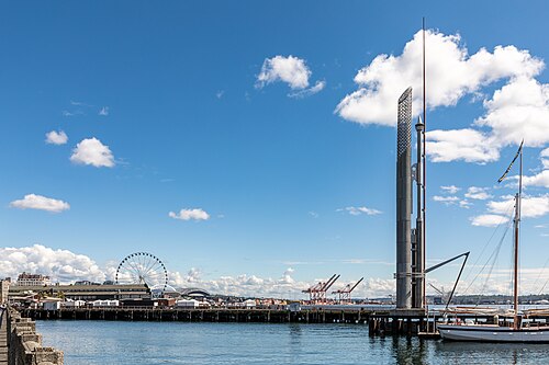 Bell Harbor Marina at the Belltown waterfront, sailboat masts at the dock, the Seattle Great Wheel and the downtown skyline across the water, a tall perforated steel sculpture standing beside the pier.