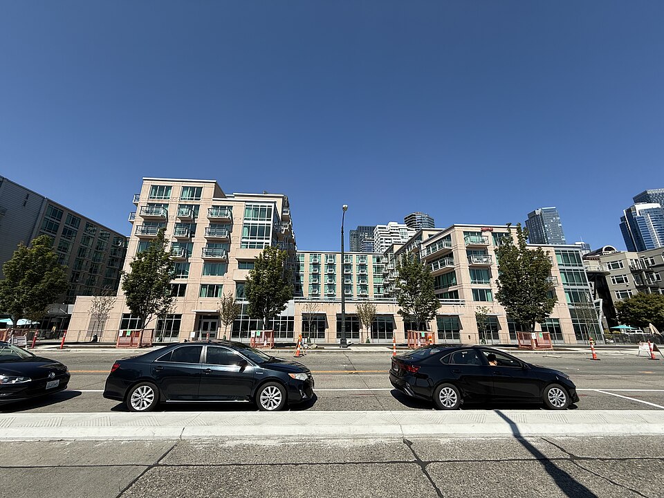 A mid rise building along Alaskan Way with tan upper floors over glass fronted ground floor retail, cars parked at the kerb, towers behind. The kind of ground floor frontage under an apartment building found along Belltown's avenues, not the nightclub itself.