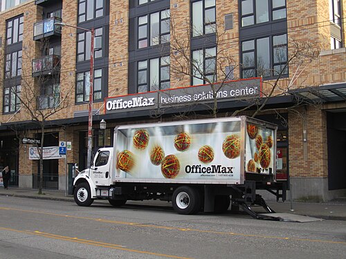 A four storey brick building on Broadway with apartment balconies above ground floor retail, a delivery truck parked at the curb and bare trees along the sidewalk.