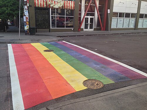 A rainbow painted crosswalk covering the intersection at 11th Avenue and Pike Street, storefronts and a red awning on the corner behind it.